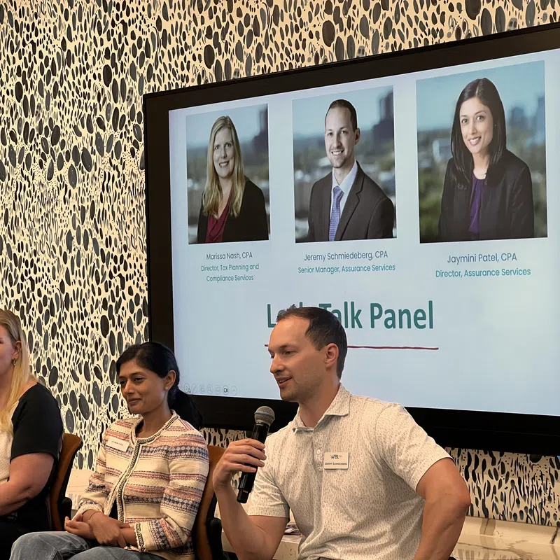 Three professionals seated for a panel discussion with a large screen showing their photos and titles behind them.