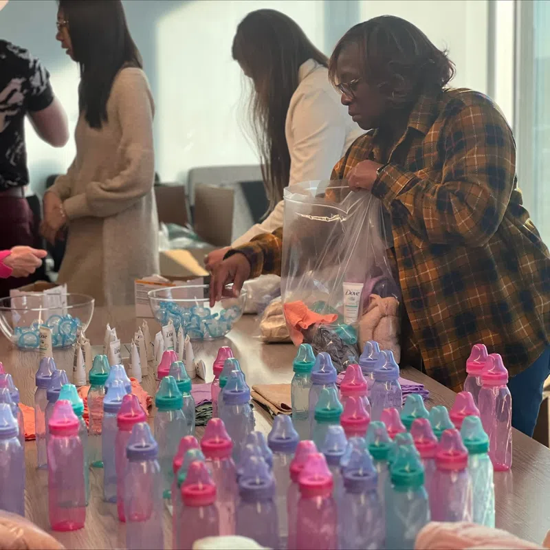 Volunteers packing baby bottles and care items into bags on a table in a bright room.