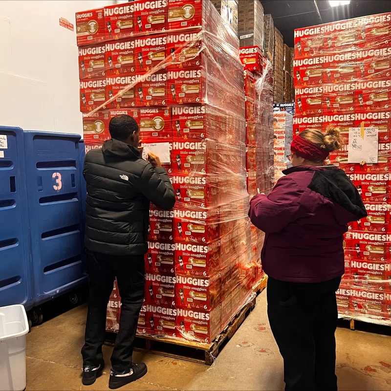 Two people in a warehouse inspecting large stacked pallets of Huggies diapers wrapped in plastic.