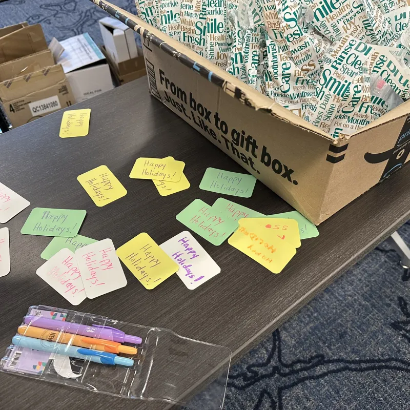 Handwritten Happy Holidays notes, markers, and a gift box filled with festive tissue paper on a table.
