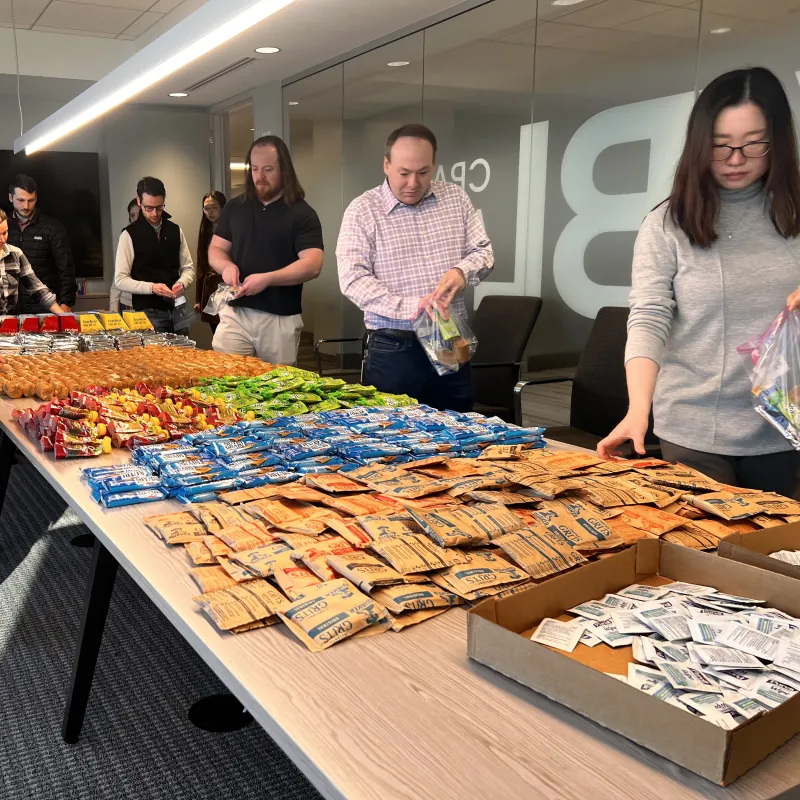 Group of people assembling snack bags at a long table with various colorful snack packages in a bright office.