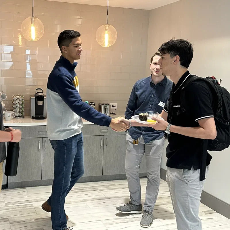 Four young men greeting and conversing in a modern kitchen area with hanging lights and coffee appliances.