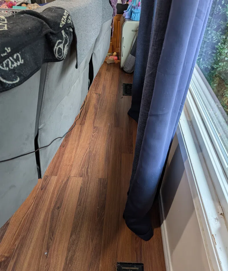 Narrow space between couch and window with wood floor, blue curtains, and visible vent on floor.