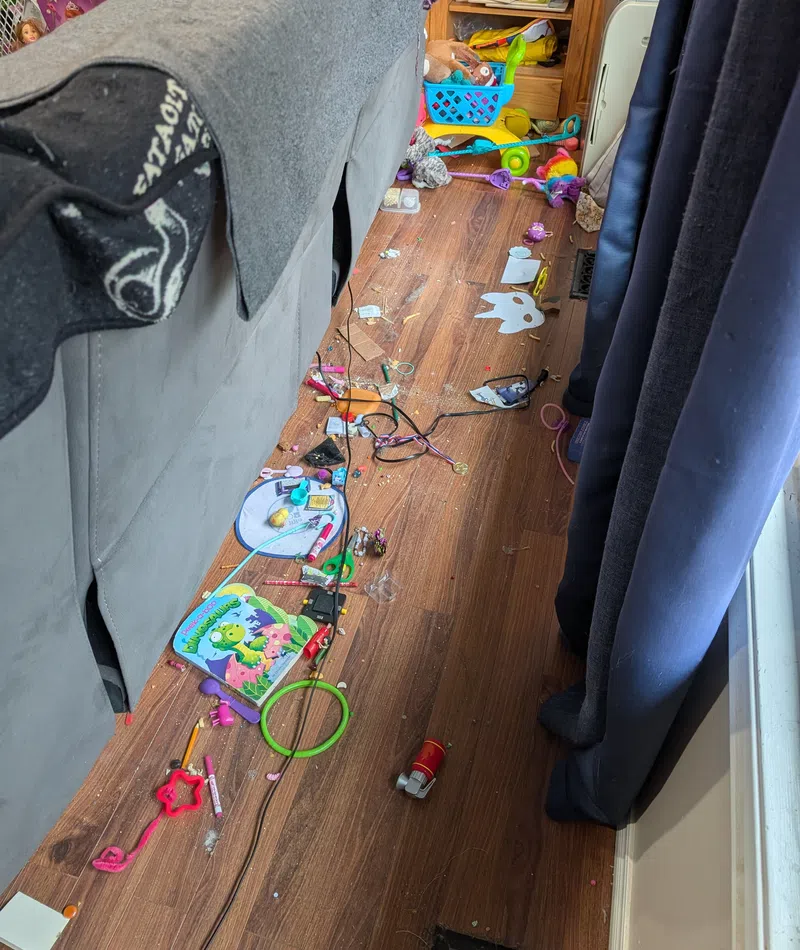 Messy living room floor with scattered children's toys, art supplies, and miscellaneous items beside a gray couch and blue curtains.