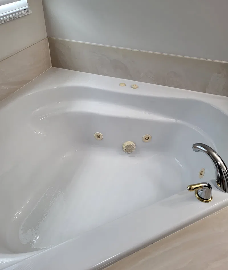 Clean white corner jetted bathtub with chrome faucet and beige marble surround in a bathroom.