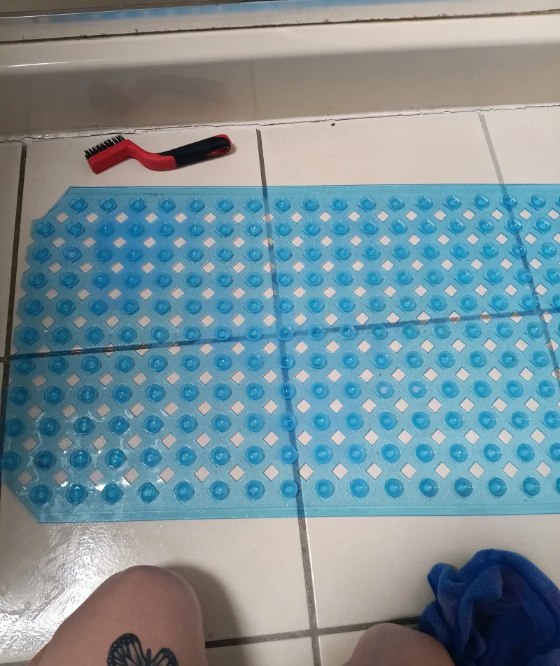 Blue bath mat with suction cups on tiled floor next to a red brush and tattooed knees.