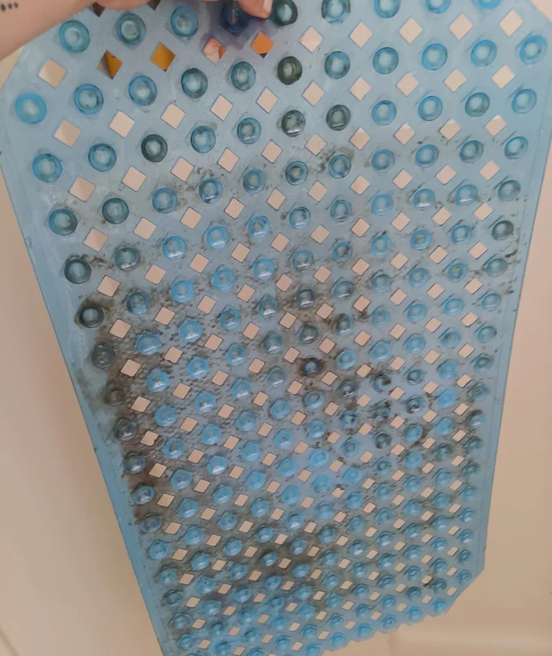 Hand holding a dirty blue rubber bath mat with suction cups against a beige background.