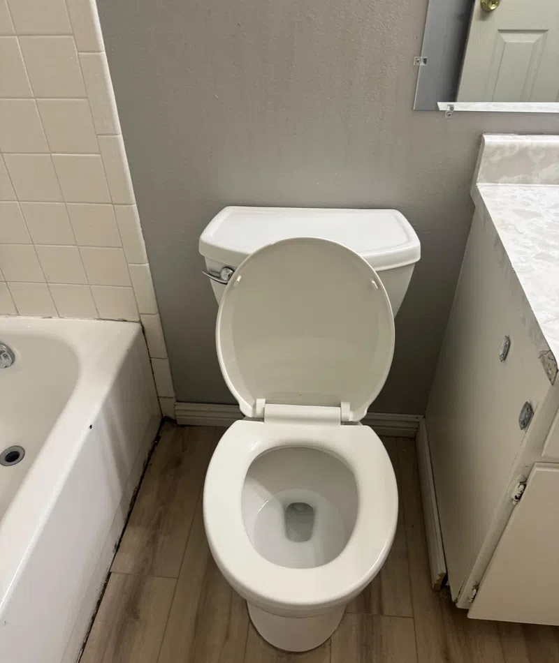 White toilet with open lid next to bathtub and white bathroom cabinet on wood floor.