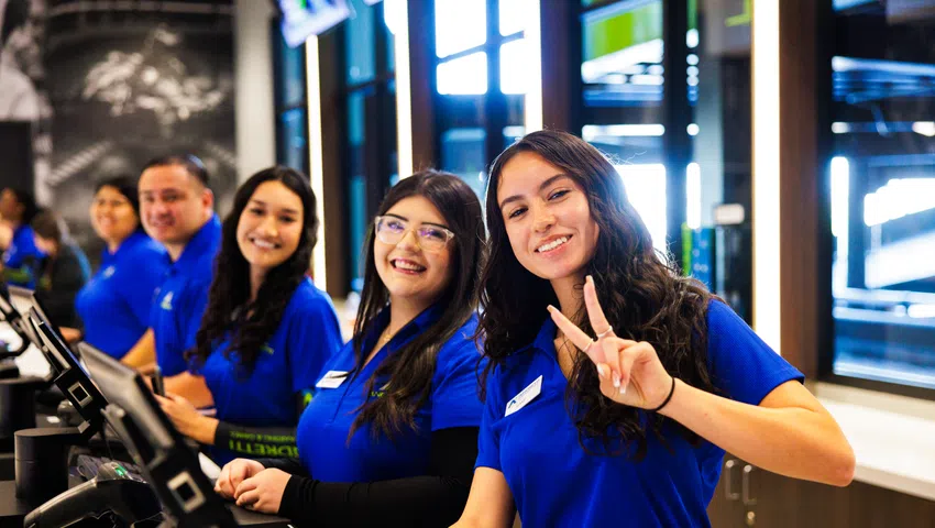 Smiling customer service team in blue uniforms at counters with computers, one woman flashing a peace sign.