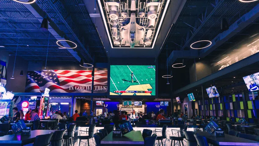 Modern sports bar interior with high tables, multiple TVs, patriotic decor, and aircraft display ceiling lighting.