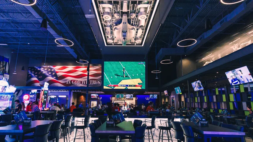 Modern sports bar interior with high tables, multiple TVs, patriotic decor, and aircraft display ceiling lighting.