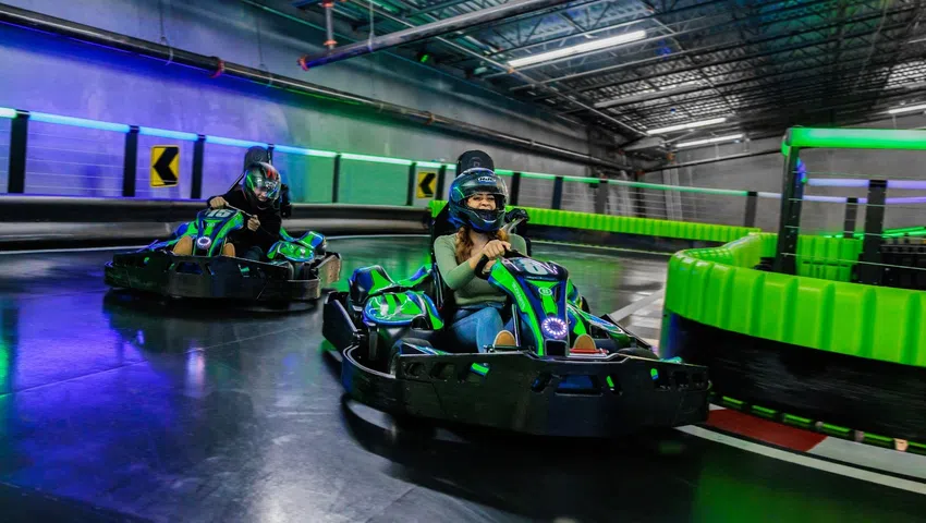 Two people racing go-karts indoors on a neon-lit track wearing helmets and smiling while driving.