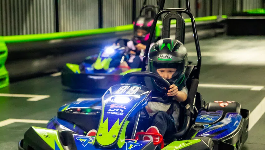 Child wearing helmet driving a blue and green go-kart on an indoor race track with another racer behind.