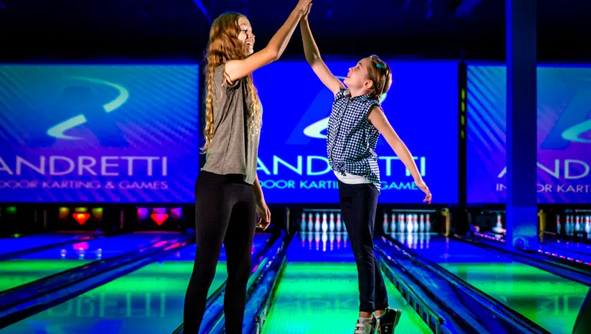 Two girls high-fiving at a colorful indoor bowling alley with Andretti signage in the background