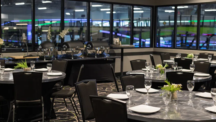 Indoor dining area with round tables set with glassware and plates, overlooking an indoor kart racing track.