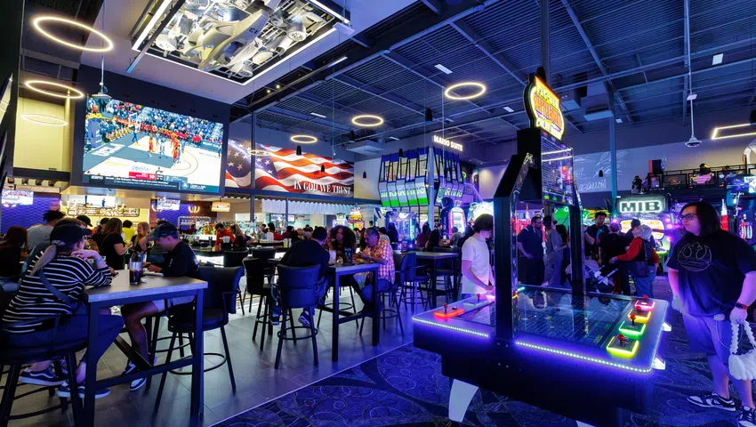 Indoor arcade and sports bar with neon lights, gaming machines, large TV showing basketball, and people socializing.