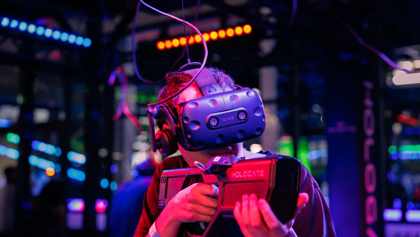 Person wearing HTC Vive VR headset and holding HOLOGATE VR gun in a colorful gaming arcade environment.