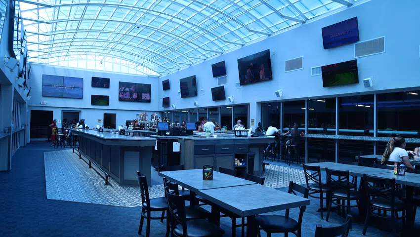 Modern sports bar interior with high glass ceiling, multiple TVs, central bar, and seating areas with tables and chairs.
