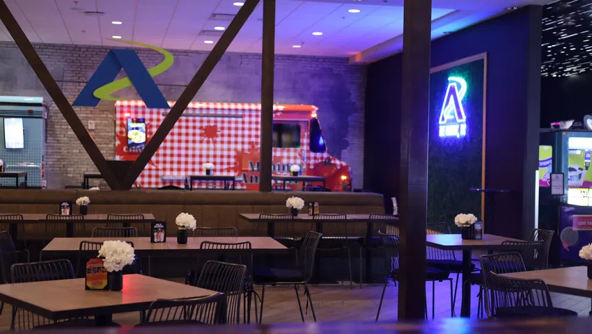 Modern cafe interior with black chairs, flower centerpieces, red checkered food truck, and neon logo on wall.