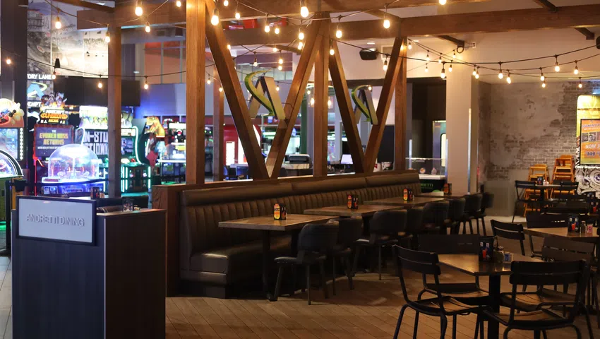 Cozy modern dining area with dark leather seating, wooden tables, string lights, and arcade games in background.
