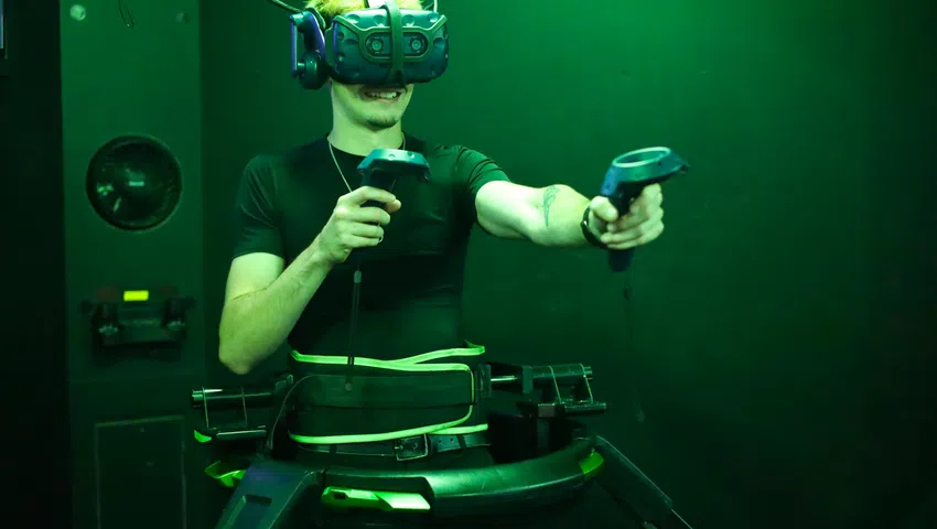Man using VR headset and controllers with motion platform in a green-lit virtual reality gaming setup.