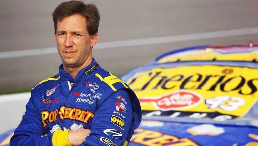 Race car driver in blue Pop Secret racing suit standing with arms crossed beside Cheerios sponsored race car.