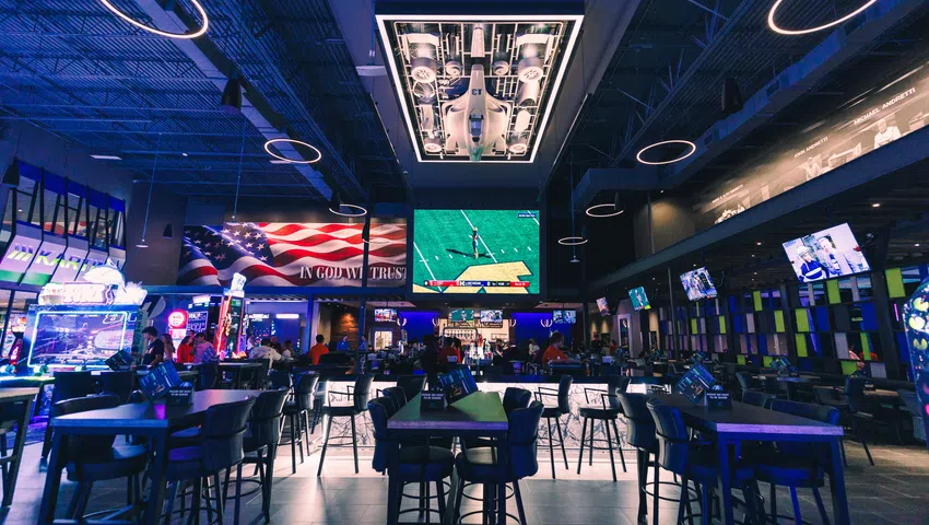 Modern sports bar interior with high tables, multiple TVs, patriotic decor, and aircraft display ceiling lighting.