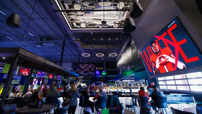 Modern sports bar interior with high tables, multiple TVs, patriotic decor, and aircraft display ceiling lighting.