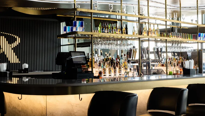 Modern bar interior with black marble counter, hanging glasses, backlit shelving, and black leather chairs.