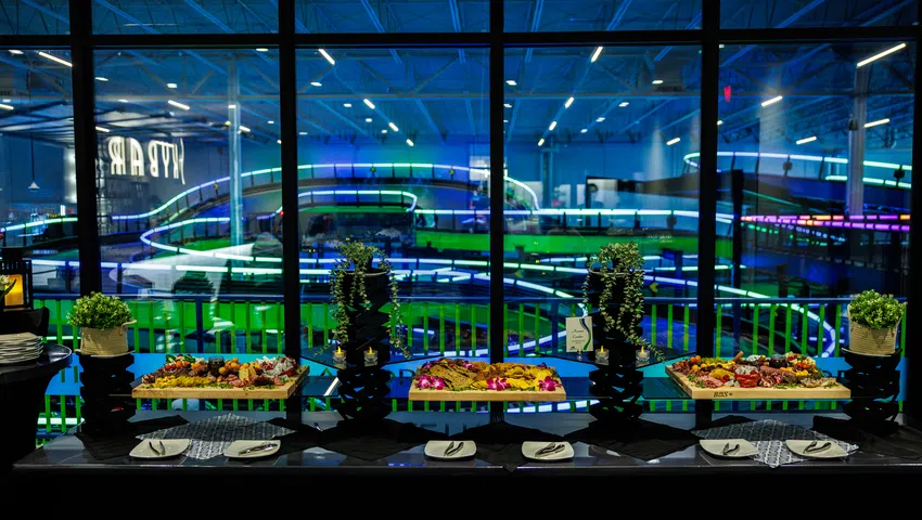 Catering buffet with assorted appetizers and greenery against neon-lit indoor go-kart track windows at night.