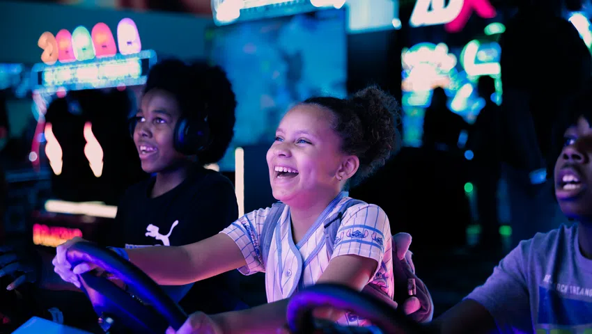 Children smiling and playing an arcade racing game in a vibrant, colorful gaming center with neon lights.