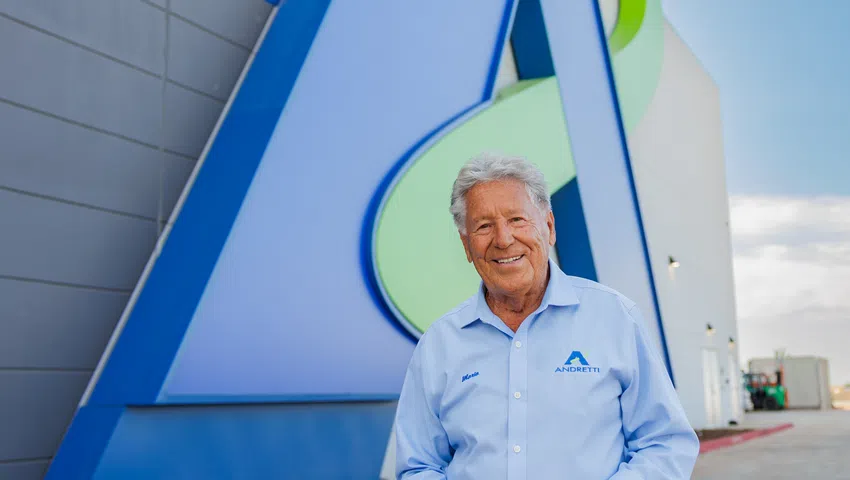 Elderly man in blue Andretti shirt stands smiling outside building with large colorful Andretti logo.