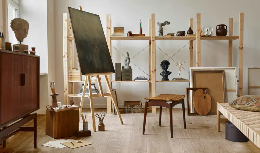 Light-filled artist studio with wooden shelves, easel, sculptures, and mid-century furniture on wood flooring
