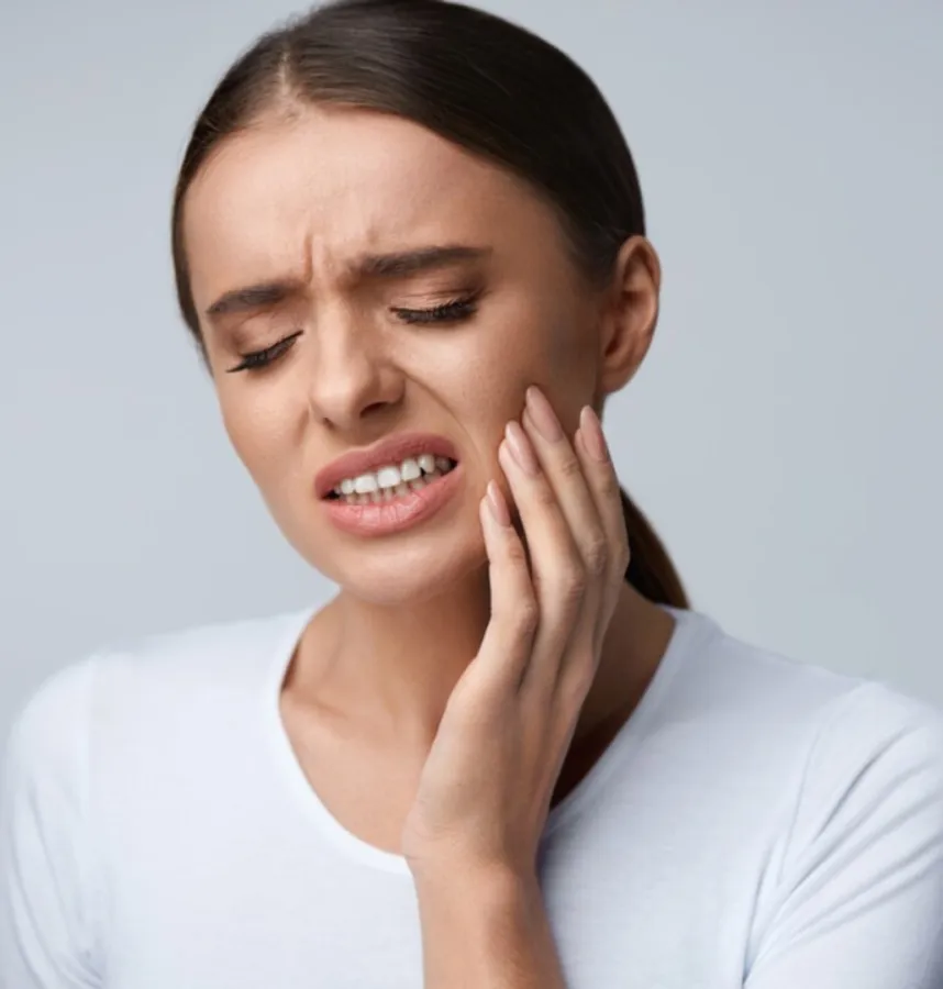Young woman in white shirt holding her cheek in pain, showing signs of toothache or dental discomfort.