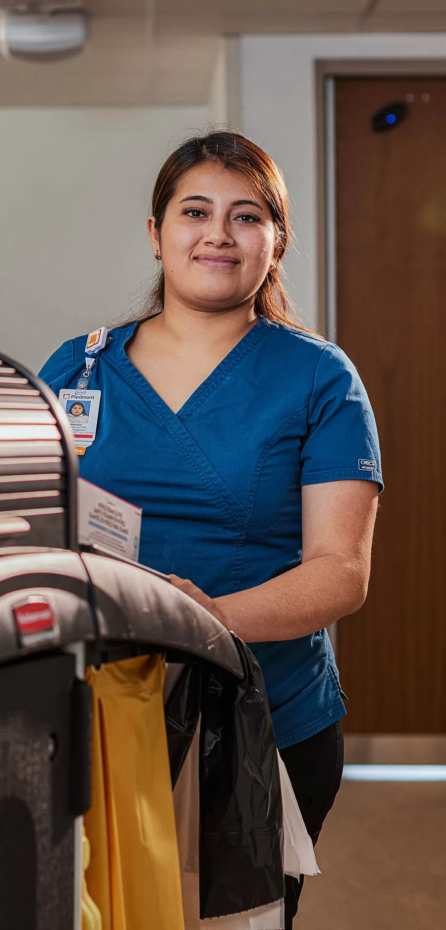 Smiling hospital janitor in blue scrubs stands beside a large cleaning cart in a healthcare hallway.