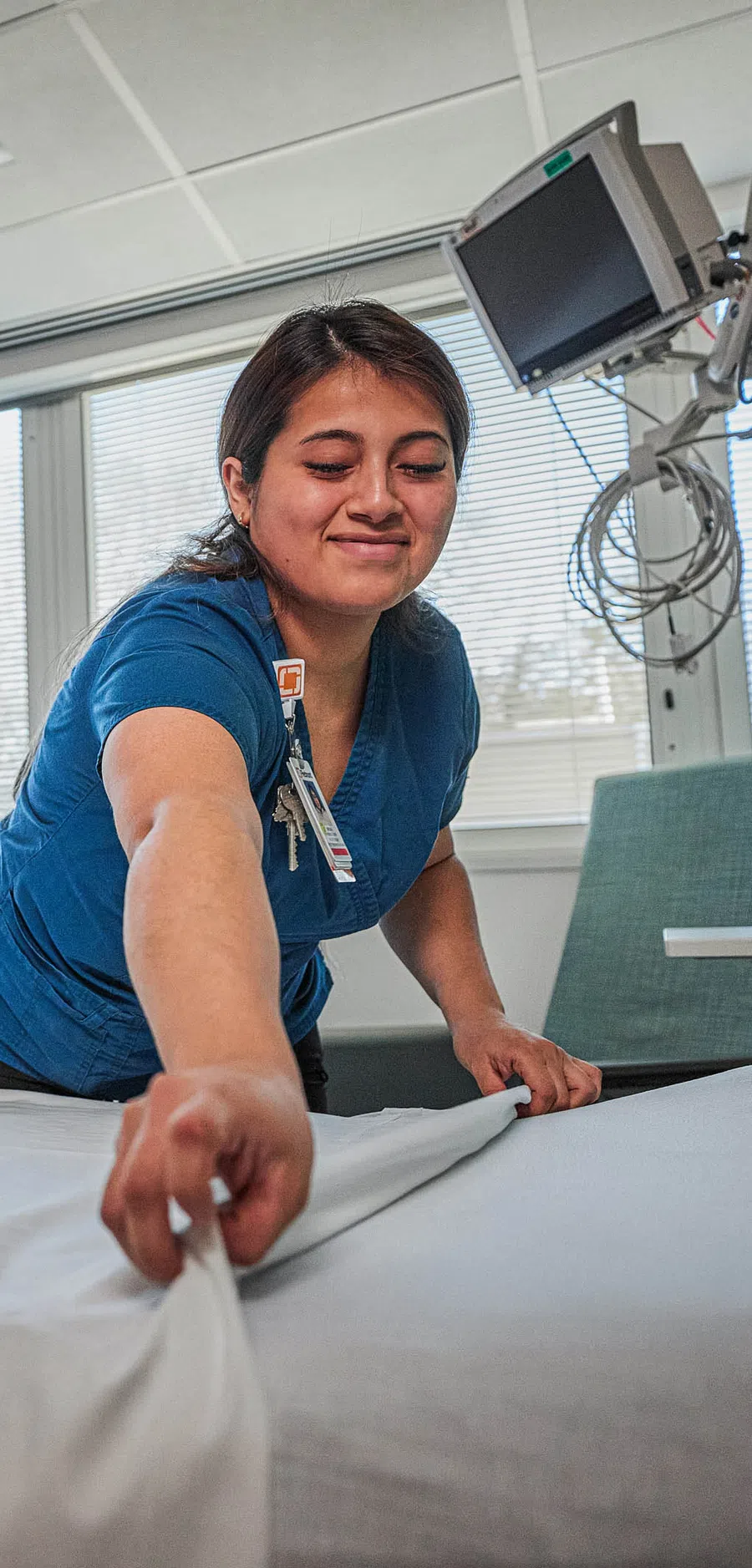 Nurse making a hospital bed in a bright patient room with medical equipment and Piedmont logo on screen.