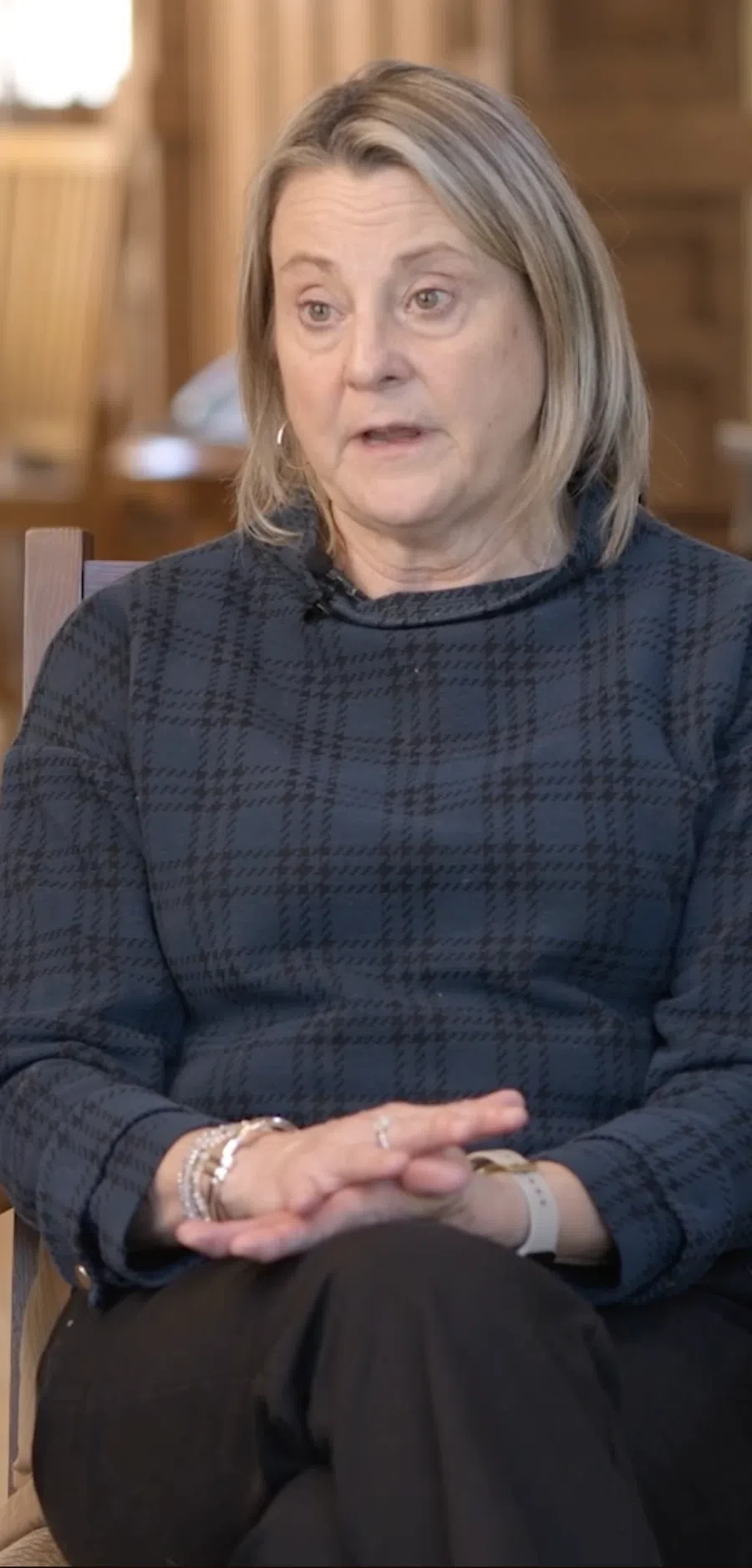 Middle-aged woman with blonde hair sitting in a wooden chair in a warm, wood-paneled room speaking thoughtfully.
