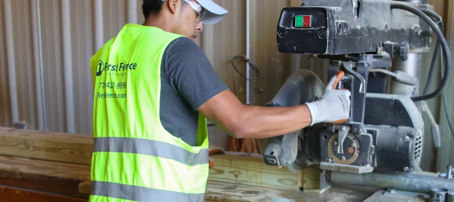 Worker wearing safety gear operating a stationary power saw to cut wood in a workshop.