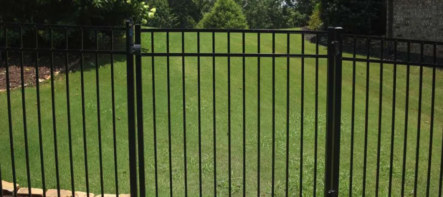 Black metal fence gate in front of green lawn with bushes and trees under a blue sky.