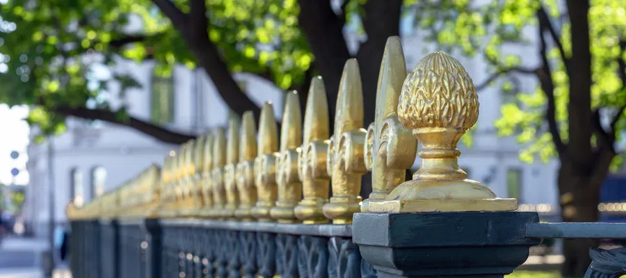 Close-up of a decorative black fence with gold accents, surrounded by lush green trees and sunlight.