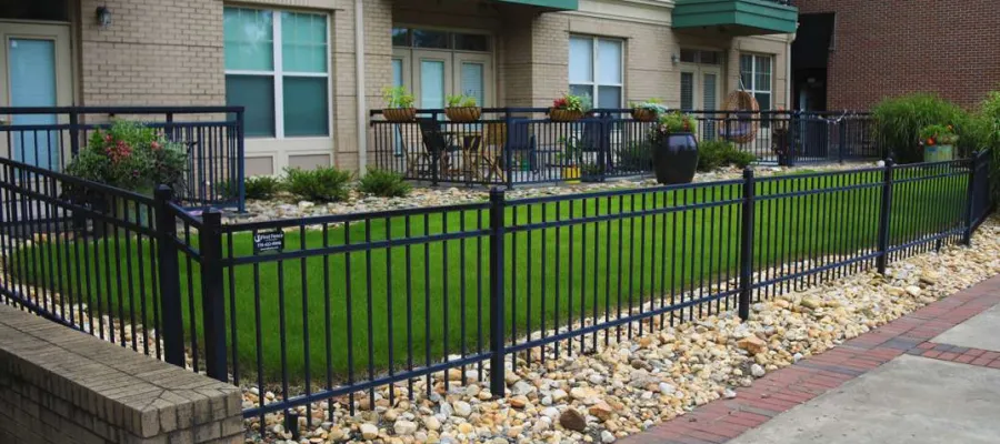 Modern apartment courtyard with black metal fence, green lawn, stone landscaping, and brick walkway.