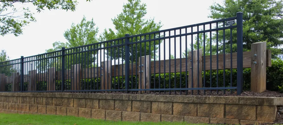 Black metal fence with vertical bars on a stone retaining wall with green grass and trees in background.