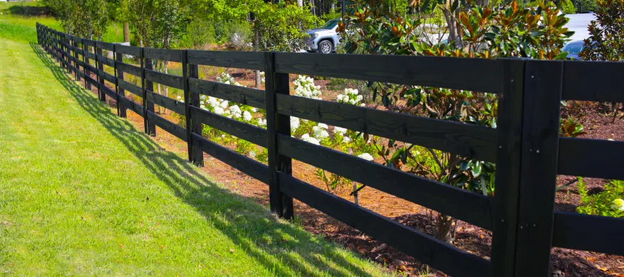 Black wooden fence casting shadows on green grass beside garden with flowering bushes and trees.