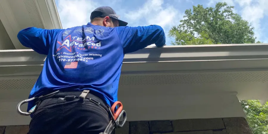 A worker in a blue shirt cleaning gutters under a clear sky with trees in the background.
