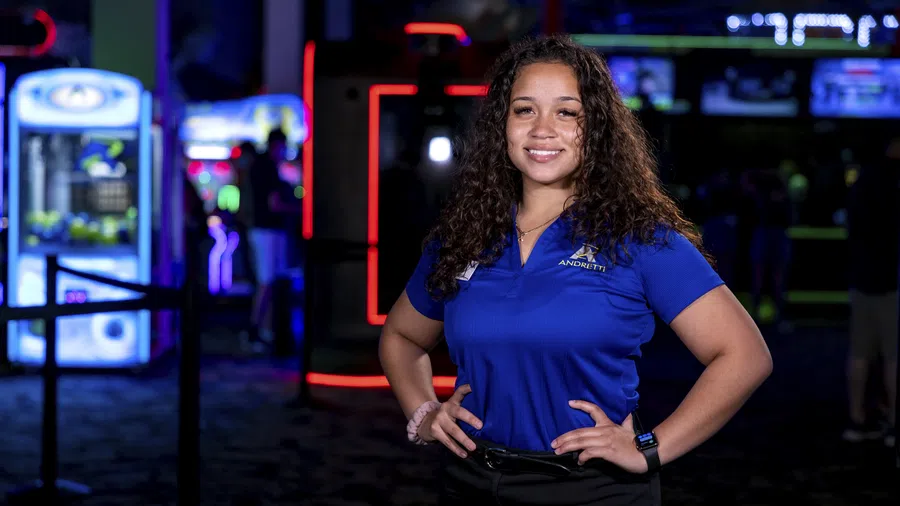 Smiling woman in a blue Andretti polo shirt stands confidently in a neon-lit arcade environment.