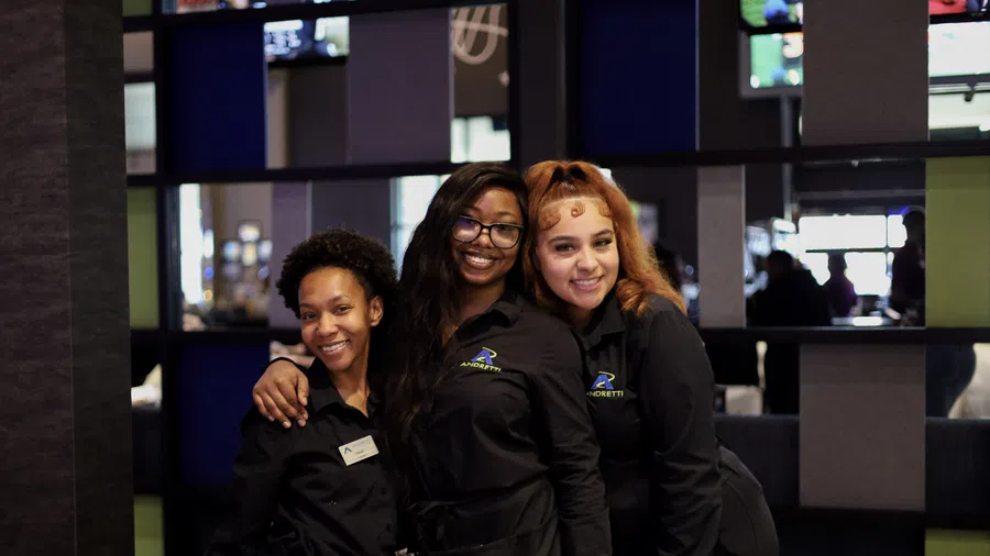 Three smiling female employees in black uniforms posing together indoors at Andretti venue.