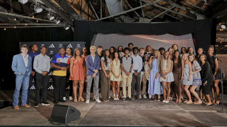 Large diverse group of young adults posing on a stage inside a rustic venue with wooden beams and lighting fixtures.