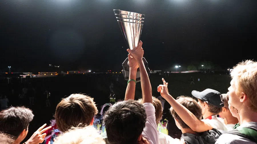 A group of young people celebrating and holding up a large silver trophy under bright stadium lights at night.