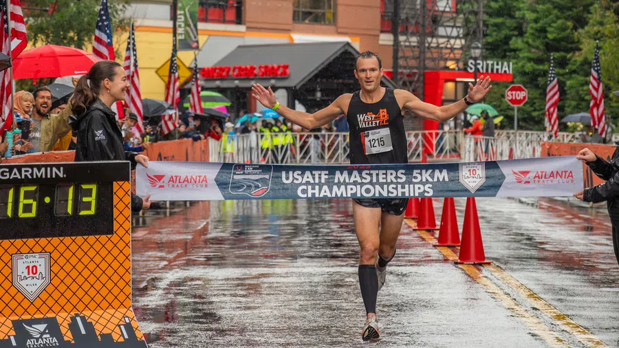 Runner crosses finish line at USATF Masters 5KM Championships with overlaid banner and rainy street scene.
