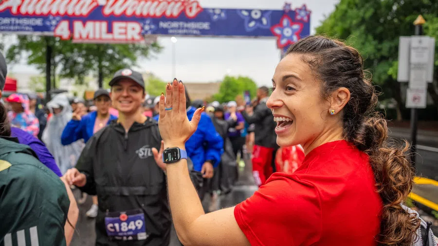Smiling woman in red shirt gives high five at Atlanta Women's 4 Miler race start line with crowd behind her.
